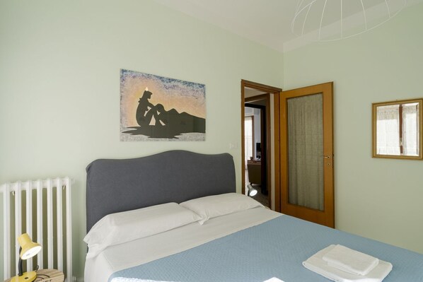 Un dormitorio con cama, lámpara de noche, un cuadro, una puerta y una ventana.
