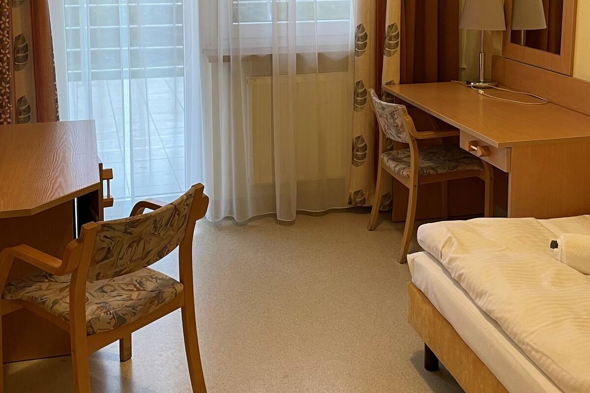 Quarto de hotel com cama, escrivaninha, cadeira, televisão e janela com cortinas.