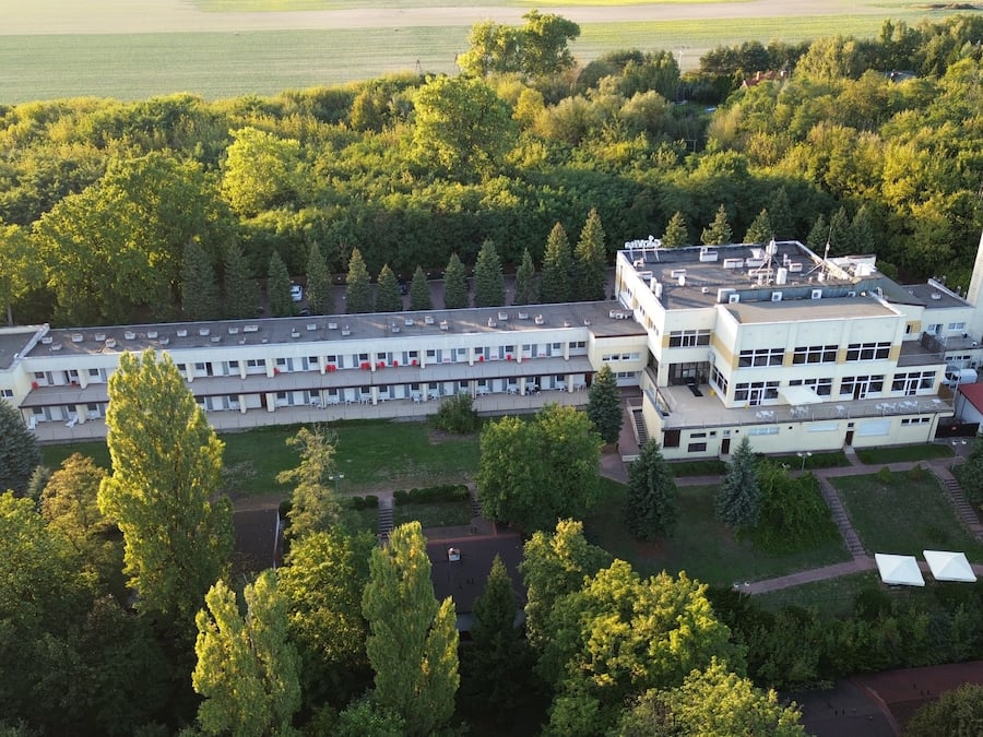 An aerial view of a large, multi-story building with a flat roof, surrounded by dense greenery.