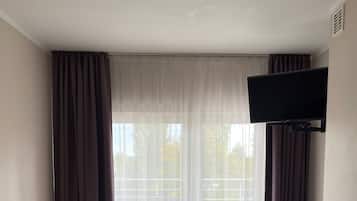 A hotel room with a single bed, a small wooden nightstand, a TV mounted on the wall, and a window with sheer curtains.
