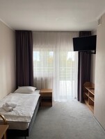Una habitación de hotel con una cama individual, una mesita de noche de madera, un televisor montado en la pared y una ventana con cortinas translúcidas.