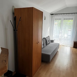 A room with a wooden wardrobe, a grey sofa, and a window with sheer curtains. - Geovita Jadwisin (Serock)