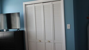Ein Zimmer mit geschlossenem weißen Kleiderschrank, einem Flachbildfernseher auf einer dunklen Kommode und einem Lichtschalter an der Wand.