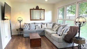 A living room with a sectional sofa, a wooden coffee table, two floor lamps, a large mirror, and a view of the outdoors through large windows.