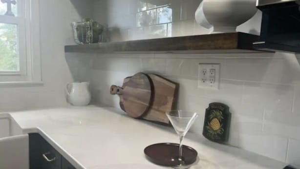 A kitchen with white countertops, a wooden cutting board, a martini glass, and a pineapple-themed wall decoration.