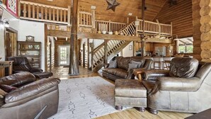 Interior de una cabaña de troncos con escalera de madera, mobiliario de cuero y decoración rústica.