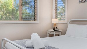 Cama bem arrumada com lençóis brancos e colcha estampada, mesa de cabeceira com abajur e janela com persianas.