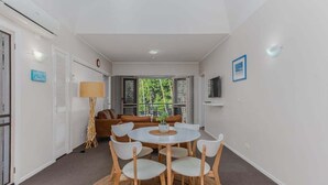 Quarto moderno com mesa de jantar redonda, cadeiras brancas, sofá marrom, TV e uma janela grande com vista para árvores.