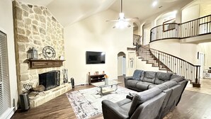A spacious living room with a stone fireplace, a large gray sectional sofa, a flat-screen TV, and a staircase leading to an upper level. - Spacious 6-bedroom house with pool and hot tub and theater room next to stadium! (Grand Prairie)