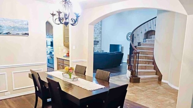 A dining area with a wooden table and chairs, a chandelier, and a staircase leading to a living area. - Spacious 6-bedroom house with pool and hot tub and theater room next to stadium! (Grand Prairie)