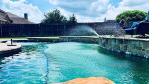 A swimming pool with a water fountain feature, surrounded by a stone deck and a wooden fence. - Spacious 6-bedroom house with pool and hot tub and theater room next to stadium! (Grand Prairie)