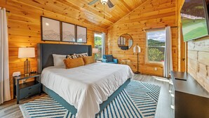 Ein Schlafzimmer in einer Holzhütte mit einem Bett, Nachttischen, einem Schreibtisch und einem großen Fenster mit Blick ins Freie.