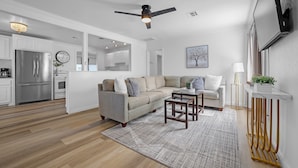 A modern living room with a sectional sofa, a coffee table, a TV mounted on the wall, and a ceiling fan. - Charming Midtown Tulsa Home Steps From Expo Square.  (Tulsa)