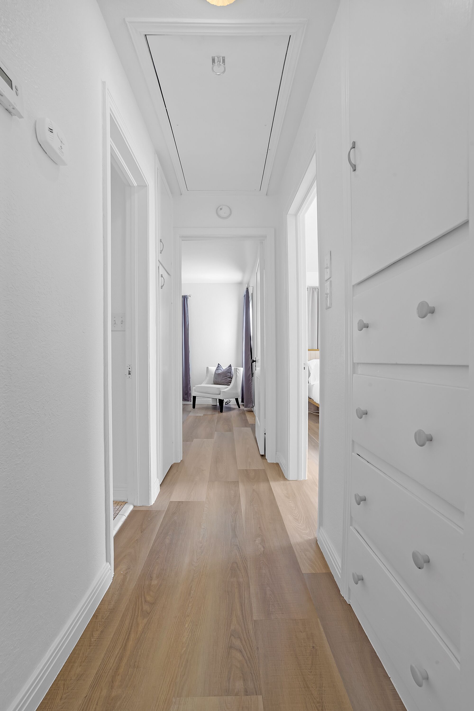 Um corredor com piso de madeira, paredes brancas e um guarda-roupa branco com várias gavetas.