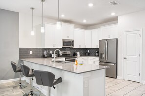 A modern kitchen with white cabinets, stainless steel appliances, and a central island with bar stools. - Epic Themed Game Room & Theater Near Disney Parks (Kissimmee)