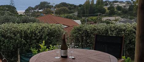 Une terrasse en bois avec une table ronde et deux chaises, offrant une vue sur un paysage côtier avec des maisons et des arbres.