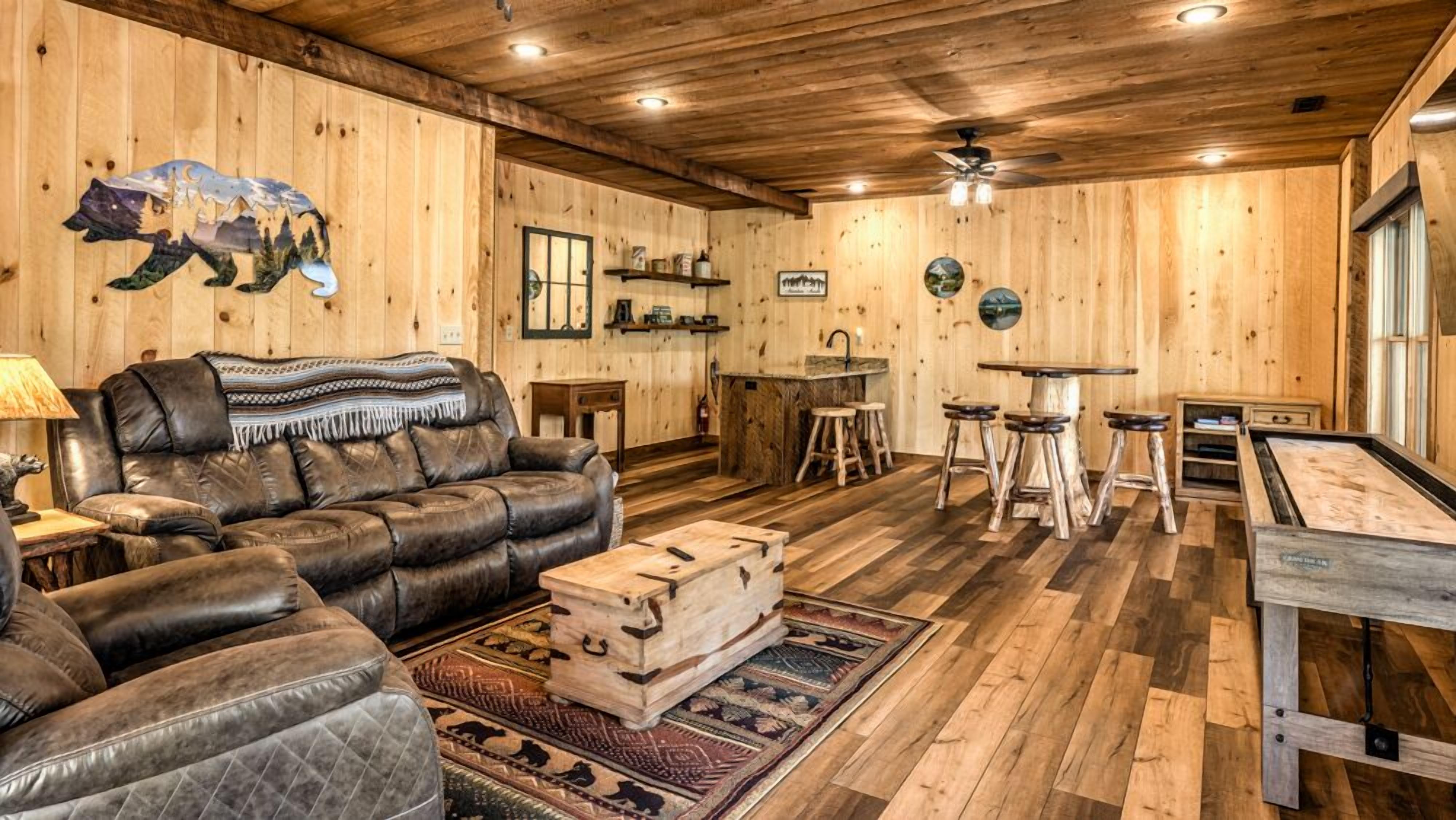 A cozy cabin interior with a leather sofa, a wooden dining table with stools, and a rustic kitchen area.