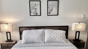 Una cama bien hecha con un edredón blanco acolchado, dos cuadros enmarcados de caballos en la pared y dos lámparas de noche sobre mesitas de madera.