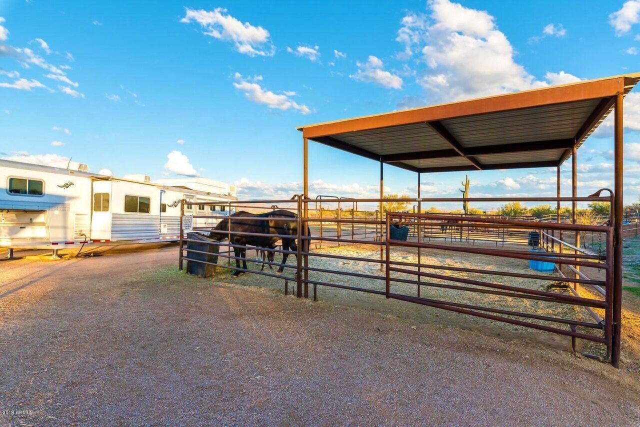 A covered outdoor area with a metal fence, a horse, and a trailer in the background.