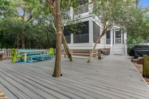 Una terraza de madera con una mesa de picnic y bancos, rodeada de árboles y un edificio blanco con balcón.