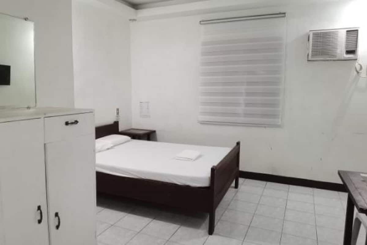 A bedroom with a bed, a bedside table, a wardrobe, a window with blinds, and an air conditioning unit.