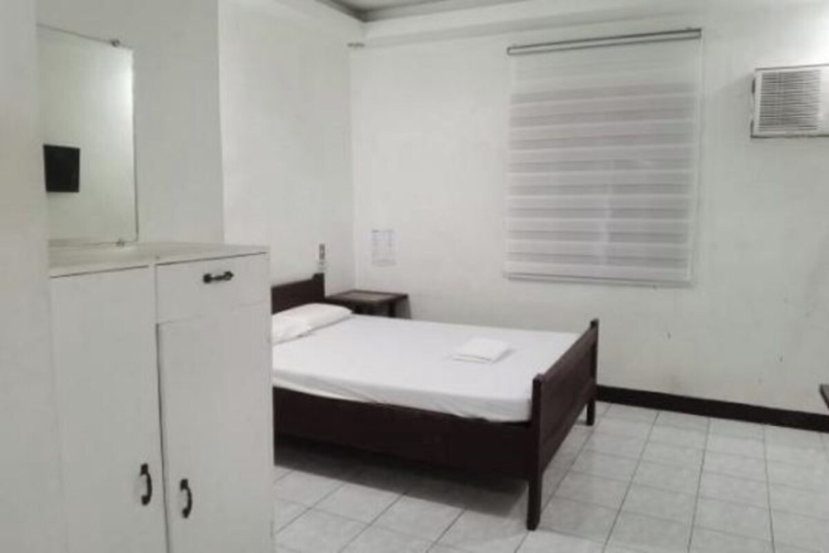 Quarto com uma cama de solteiro, mesa de cabeceira, espelho e janela com persianas.