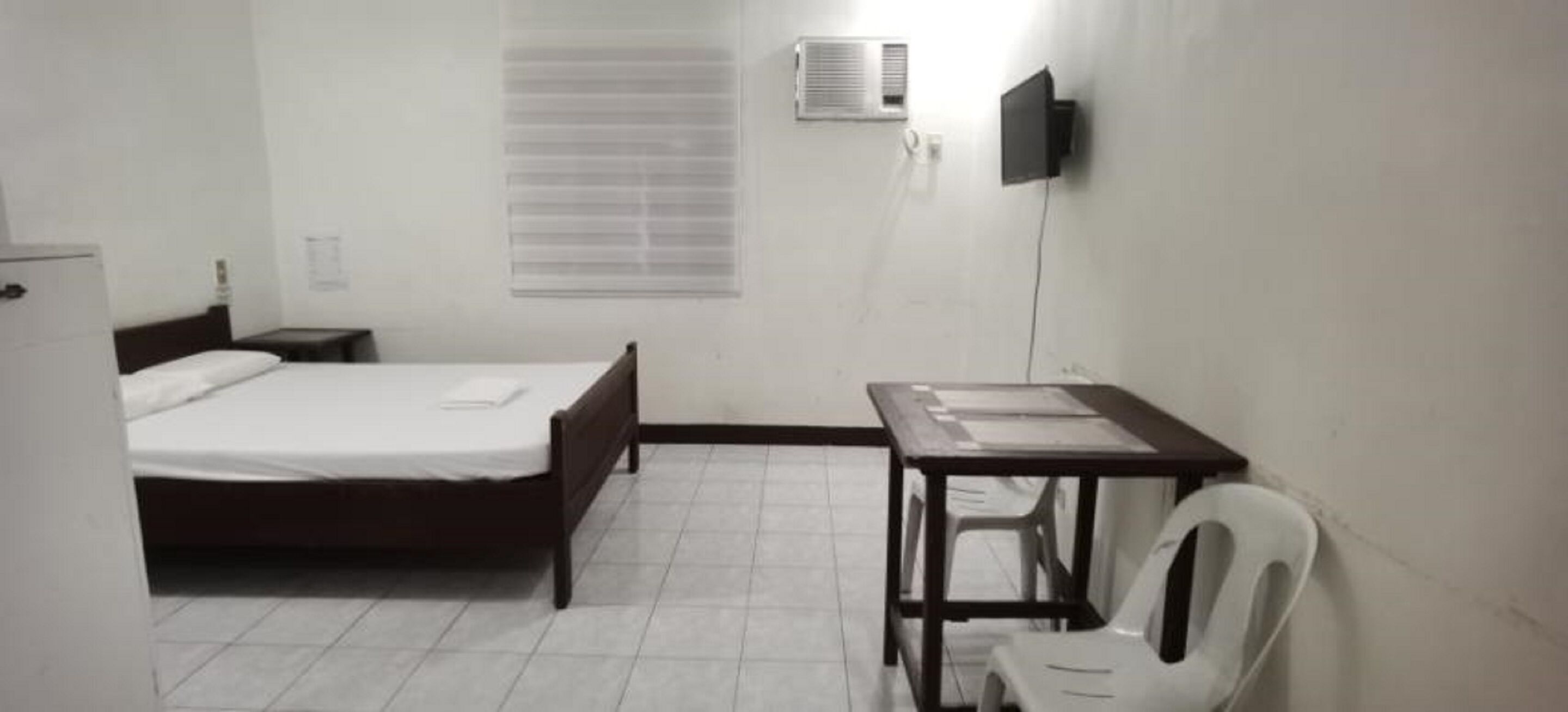 A small, sparsely furnished room with a bed, a desk, a chair, and a wall-mounted air conditioning unit.