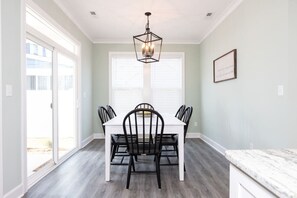 Un coin repas avec une table blanche et des chaises noires, un lustre et une fenêtre avec des stores.