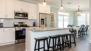 A modern kitchen with white cabinets, stainless steel appliances, a central island with bar stools, and pendant lighting. - Coastal Catch: Walk to Boardwalk (Virginia Beach)