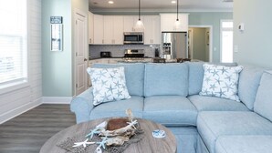 A modern living room with a light blue sofa, a round coffee table with decorative items, and a kitchen in the background. - Coastal Catch: Walk to Boardwalk (Virginia Beach)