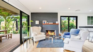 Una sala de estar moderna con chimenea, una puerta de cristal que da a un patio, y una cocina visible a través del cristal.