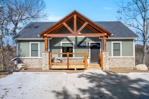 A two-story house with a stone foundation, a wooden front porch, and a garage door. - Private Hot Tub, Expansive Decks & Taneycomo Views in Charming Farmhouse with Free Tickets (Hollister)