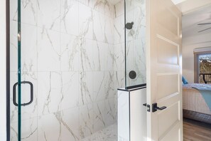 A room with a shower area featuring white marble tiles, a glass door, and a bed with a blue pillow. - Private Hot Tub, Expansive Decks & Taneycomo Views in Charming Farmhouse with Free Tickets (Hollister)