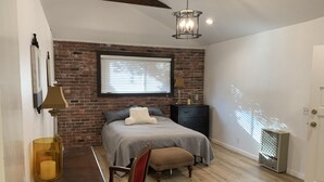 Un dormitorio con pared de ladrillo, cama con colcha gris, escritorio de madera con lámpara, silla y alfombra estampada.