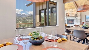 Ein Essbereich mit einem Tisch für vier Personen, einer Obstschale und Weingläsern. Durch große Fenster ist eine Berglandschaft zu sehen.