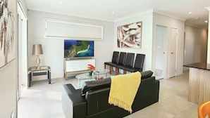 A modern living room with a black sofa, a glass coffee table, a TV, and a painting on the wall. - Delightful 2-level home in Bardia (Bardia)