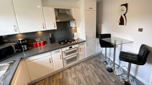 A compact kitchen with white cabinets, a gas stove, and a coffee maker. There is a small dining area with a glass table and two black chairs. - NEW Super 2 Bedroom Flat in Falkirk (Falkirk)