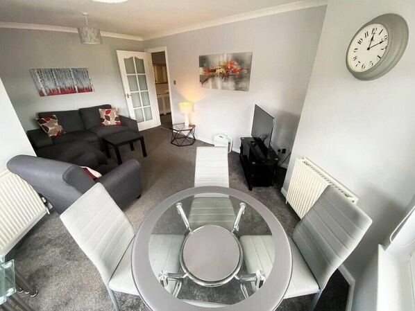 A modern living room with a round glass table, grey sofas, a TV stand, and a clock on the wall.
