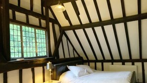 Een slaapkamer met een groot raam, een bed met witte lakens, een nachtkastje met een lamp en zichtbare houten balken aan het plafond.