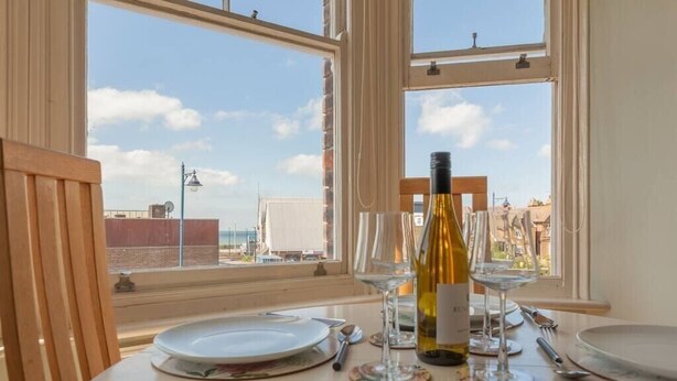 Une table dressée pour un dîner à deux, avec des verres à vin et une bouteille de vin, donnant sur un paysage urbain à travers de grandes fenêtres.