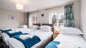A hotel room with three beds, each with white linens and blue blankets. There is a window with curtains, a bedside table with a lamp, and a wall-mounted heart decoration. - Stunning 3 Bedroom Home Hillsborough Sheffield (Hillsborough)