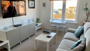 A modern living room with a sofa, coffee table, and a TV mounted on the wall. There is a window with a view of buildings outside. - Superb One Bedroom Getaway in Dysart Kirkcaldy (Dysart)