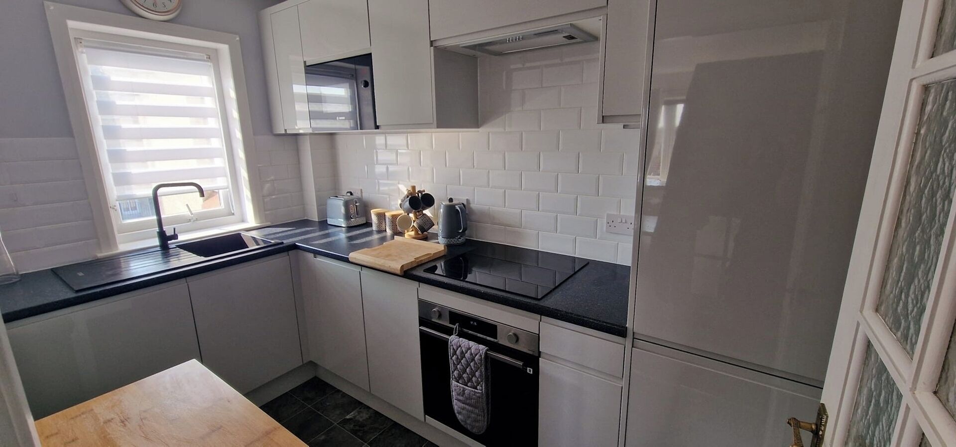 A kitchen with white cabinets, a black countertop, a window with blinds, a toaster, a kettle, and a dishwasher.