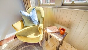 Un fauteuil jaune avec un coussin bleu, une petite table d’appoint en bois sur laquelle se trouvent un livre et une tasse, et une fenêtre donnant sur des immeubles.