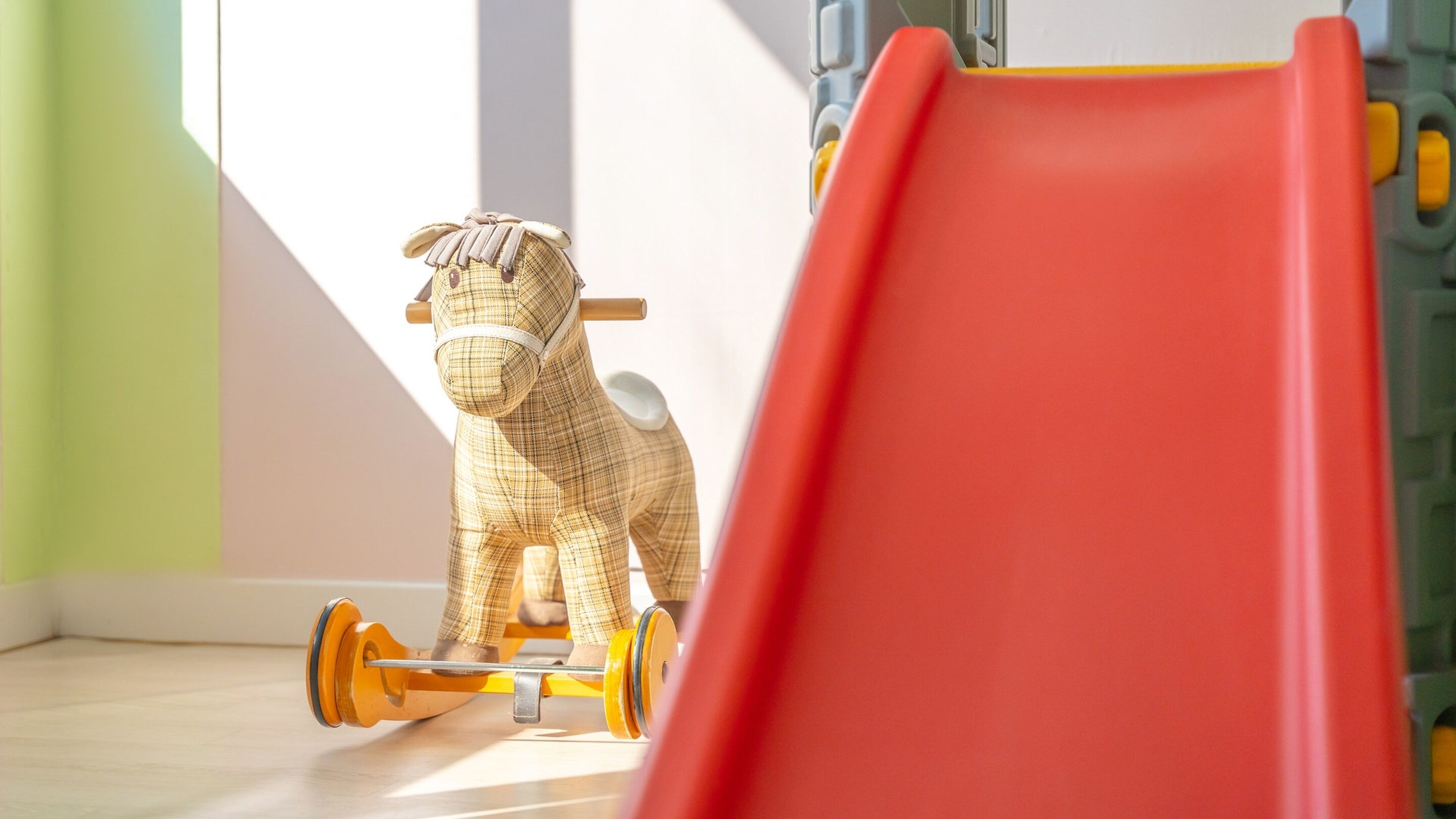 Un cheval en jouet sur une petite voiture jaune et orange, un toboggan rouge et un mur vert.