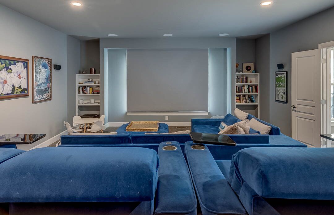 A modern living room with a large flat-screen TV, blue sofas, white chairs, and a bookshelf.