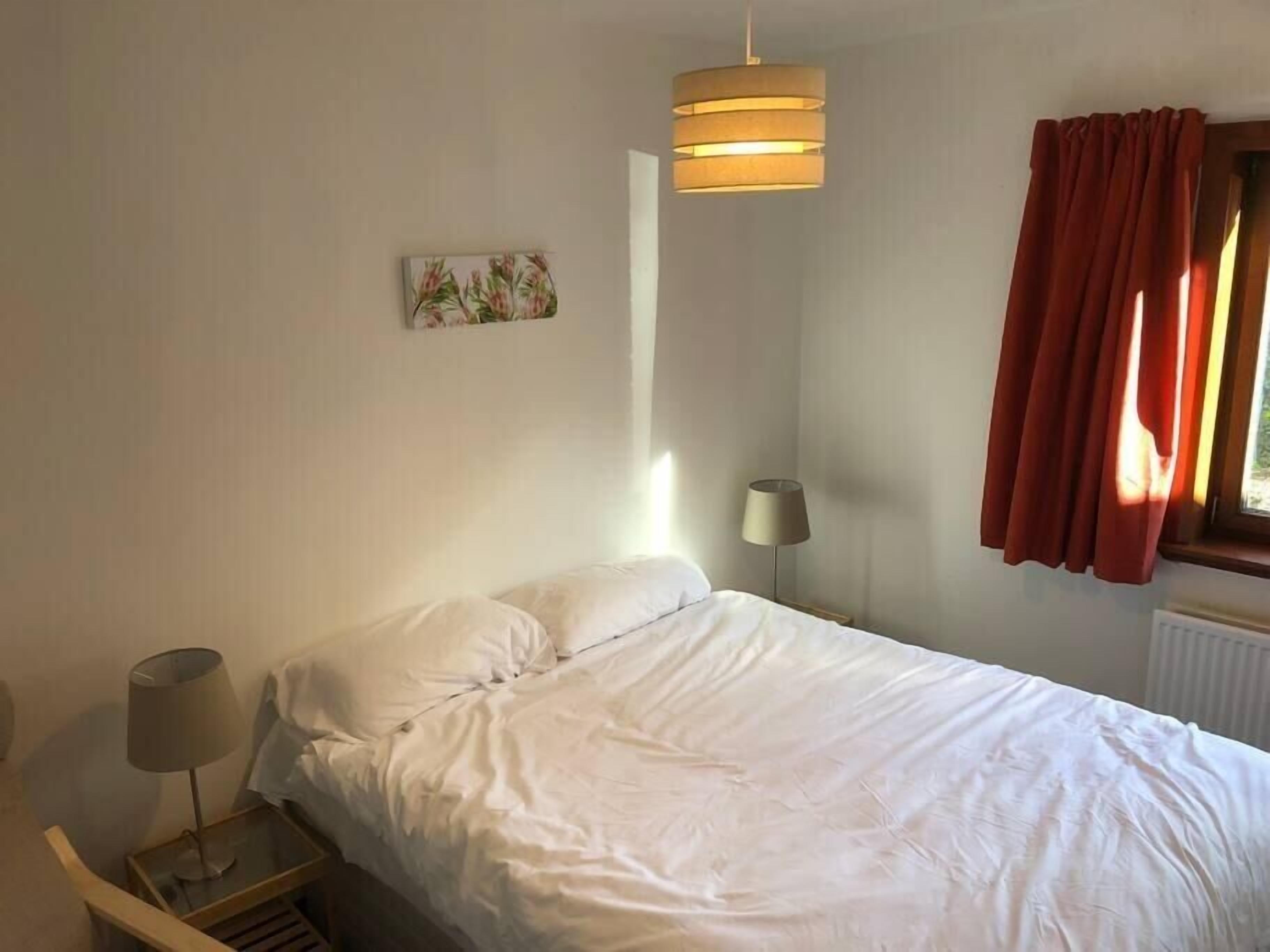 Une chambre à coucher avec un lit, une lampe de chevet, un luminaire suspendu, une fenêtre avec des rideaux rouges et un tableau accroché au mur.