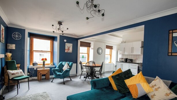 A living room with teal sofas, a dining area, and a kitchen visible through an open doorway. - Charming 2 Bedroom Flat in the Heart of Kirriemuir (Angus)