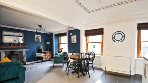 A spacious living room with a fireplace, a dining area with a round table and chairs, and a kitchen area with a radiator and window. - Charming 2 Bedroom Flat in the Heart of Kirriemuir (Angus)