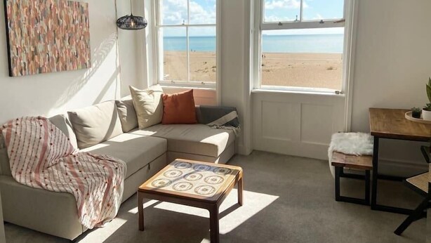 Ett mysigt vardagsrum med en beige soffa, ett litet träbord och två fönster med utsikt över en strand.